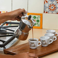 Person pouring coffee from a silver espresso pot into white mugs with 'Cafe La Llave' branding on a wooden surface.